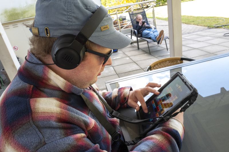 Osoba w słuchawkach i czapce używa tabletu na tarasie. W tle ktoś siedzi na leżaku, patrząc w telefon.